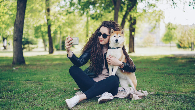 Cheerful blogger loving pet owner is taking selfie with her dog using smartphone, human and animal are sitting on lawn in the park and posing, woman is caressing and kissing dog.