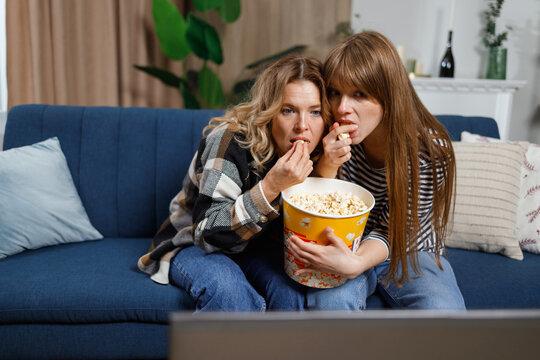 Middle age woman and her adult daughter are excitedly watching scary movie on laptop sitting on the sofa in living room