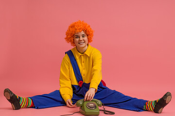 a funny clown in a wig and a yellow-blue suit sits with his legs apart and puts down the receiver from a retro phone on a colored background