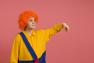cheerful girl in a clown costume and a bright wig points down on a colored background
