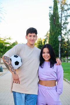 Two young latin friends posing with a soccer ball and smiling at street in a park.