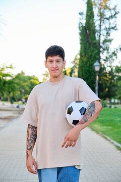 An Ecuadorian young man posing with a soccer ball and smiling at street in a park.