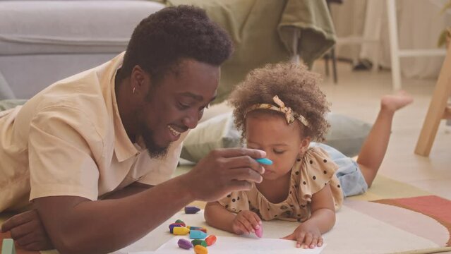 Young Black man with toddler daughter lying together on rug in living room and drawing with colorful waxed crayons