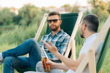 Two men sitting in camping chairs drinking beer and chatting outdoors.