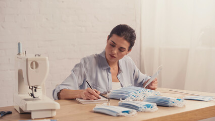 Seamstress holding medical mask and writing on notebook near sewing machine.