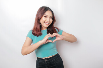 An attractive young Asian woman wearing a blue t-shirt feels happy and a romantic shapes heart gesture expresses tender feelings