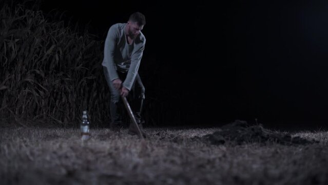 Killer digs grave at night. Young scary man digs hole in field. Long shot.