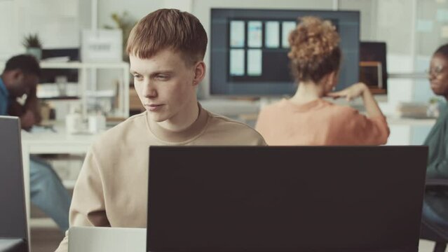 Young Caucasian male programmer looking at computer monitor while working in IT office with diverse colleagues