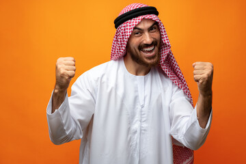 Successful smiling young Arab man celebrating victory on orange background