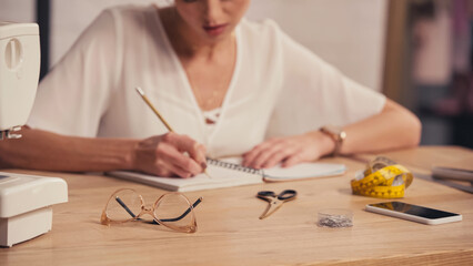Cropped view of eyeglasses near sewing machine and blurred designer writing on notebook in atelier.