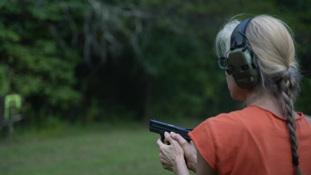 Mature, elderly woman fires a handgun at a shooting range target outside on a summer day.