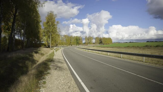 Gray asphalt winding road in autumn. Protective barriers on the side of the road. Yellow birches and bushes against the background of olak trees against a blue sky. Warm autumn day in Latvia