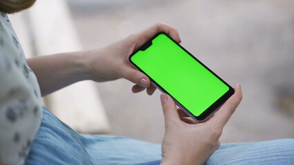 The girl holds a smartphone horizontally and watches the video on the screen. Green screen smartphone for tracking. Concept - social networking, mobile Internet, media content on a smartphone