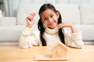 Asian little girl is putting the coins to money box and looking to camera with smile. Concept of kid education about financial, saving, planing, economy and business.