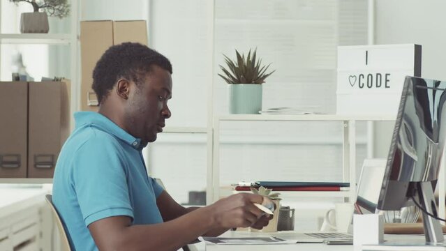 Young African American male programmer in blue polo shirt taking notes on post its and using laptop while working in IT office