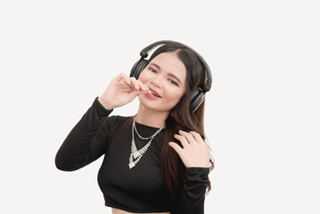 Fototapeta premium A cute Asian female eating lollipop listens to music with her wireless headphones while posing for a studio photo with white bacground.