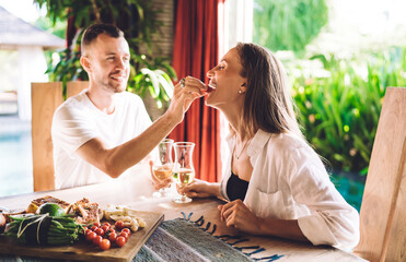 Cheerful female enjoying spending time with Caucasian boyfriend talking and eating tasty food at terrace, blurred husband feeding wife during positive date time feeling romantic togetherness