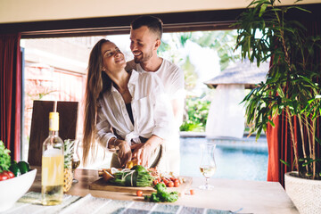 Excited male and female with fresh vegetables laughing while cooking brunch meal at house terrace, joyful Caucaisan marriage with food smiling and rejoicing togetherness in homey interior