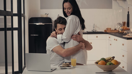 sexy woman hugging happy man near breakfast and laptop in kitchen.