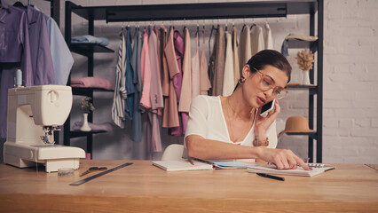 Designer talking on smartphone near notebooks and sewing machine in atelier.