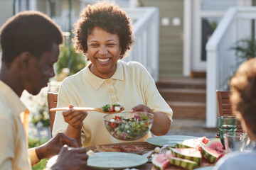 Portrait of smiling African American woman serving food to family while enjoying dinner together outdoors in sunlight - Powered by Adobe