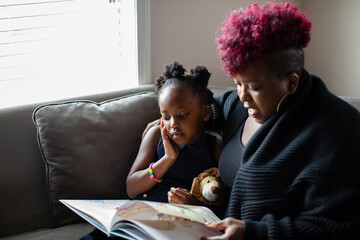 African American mother and daughter reading book together on couch 