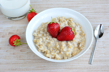 cooked oat flakes in white bowl decorated with strawberry, teaspoon and glass of milk