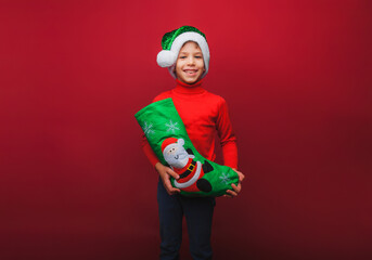 a boy in a knitted red sweater and a Santa Claus hat holds a Christmas boot for gifts. Cute baby is waiting for Christmas and a new kind. insulation on a red background.
