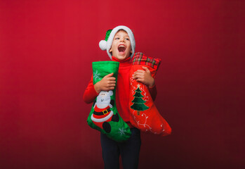 a boy in a knitted red sweater and a Santa Claus hat holds a Christmas boot for gifts. Cute baby is waiting for Christmas and a new kind. insulation on a red background.