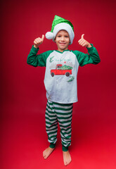 a boy in a Santa hat and New Year's pajamas shows a cool gesture isolated on a red background. the concept of Christmas and new year