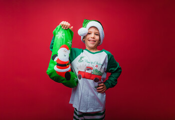Caucasian little boy in New Year's pajamas and Santa Claus hat stands with a bag of New Year's gifts, slyly looks at the camera, copy space, red isolated background © inna717