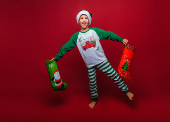 Caucasian little boy in New Year's pajamas and Santa Claus hat stands with a bag of New Year's gifts, slyly looks at the camera, copy space, red isolated background