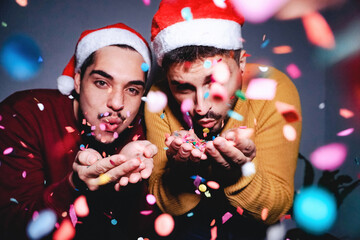 Happy gay men couple having fun blowing confetti wearing Santa Clause hats during Christmas eve - Focus on hands