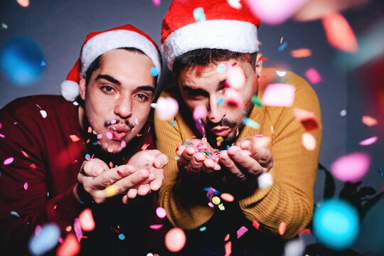 Happy gay men couple having fun blowing confetti wearing Santa Clause hats during Christmas eve - Focus on hands
