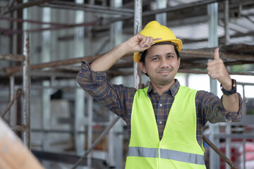 portrait of asian male construction worker hands on safety helmet and thumbs up sign and smiles cheerfully at construction site