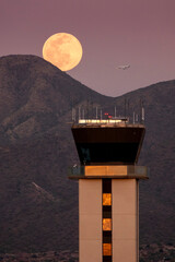 This large full moor rises over the McDowell Mountaind range and the airport tower at Scottsdale, Arizona Airport. A business jet on approach to the airport is visible close to the full moon. - 538936638