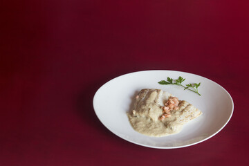 Plating of white fish with green sauce on an elegant plate garnished with a sprig of fresh parsley on a red textured studio background with copy space. Easy and healthy recipes.