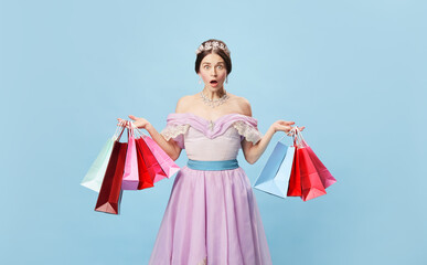 Shocked princess. Young woman in image of young queen holding bright shopping packages looking at camera with surprise isolated on blue background.