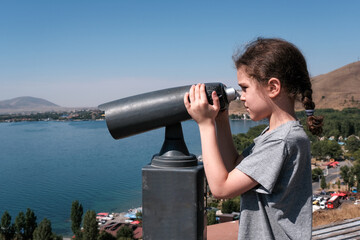Fototapeta na wymiar 10-year-old boy looks through coin operated binocular (binoscope) from Sevanavank monastery on sunny summer day. Sevan lake, Armenia.