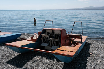 Beach on Sevan lake with old paddle boat (pedalo, water bike) on pebble and girl in the water on sunny summer day, Armenia.