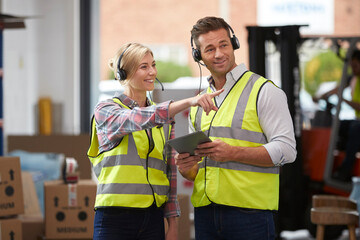 Male And Female Workers Wearing Headsets In Logistics Distribution Warehouse Using Digital Tablet