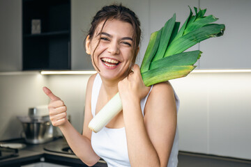 Cheerful young woman in the kitchen holds a leek in her hands.