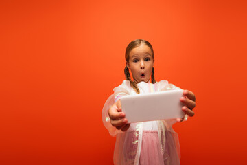girl with open mouth and thrilled face expression taking selfie on cellphone isolated on orange