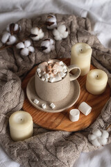Cozy home still life: a cup of hot chocolate or cocoa with marshmallows on a wooden board, a wool sweater, candles and a sprig of cotton.