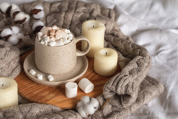 Obraz na płótnie Canvas Cozy home still life: hot chocolate or cocoa with marshmallows on a wooden board, a wool sweater, candles and a sprig of cotton. Horizontal photo. A concept for wishing a good day. Selective focus.