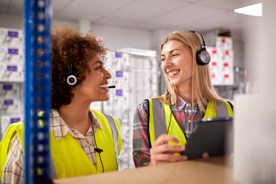 Two Female Workers Wearing Headsets In Logistics Distribution Warehouse Using Digital Tablet