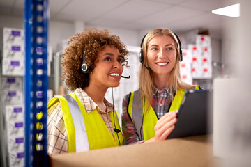 Two Female Workers Wearing Headsets In Logistics Distribution Warehouse Using Digital Tablet