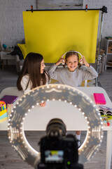Obraz na płótnie Canvas happy video blogger putting on wireless headphones near friend and blurred digital camera with circle light