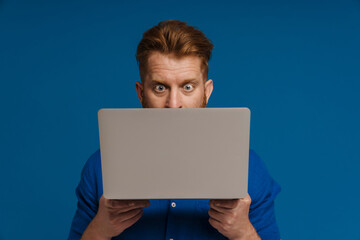 Ginger shocked man looking at camera while posing with laptop