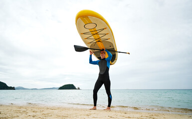 An athletic man in a wetsuit carries a SUP board on his head along the sandy seashore. Extreme...